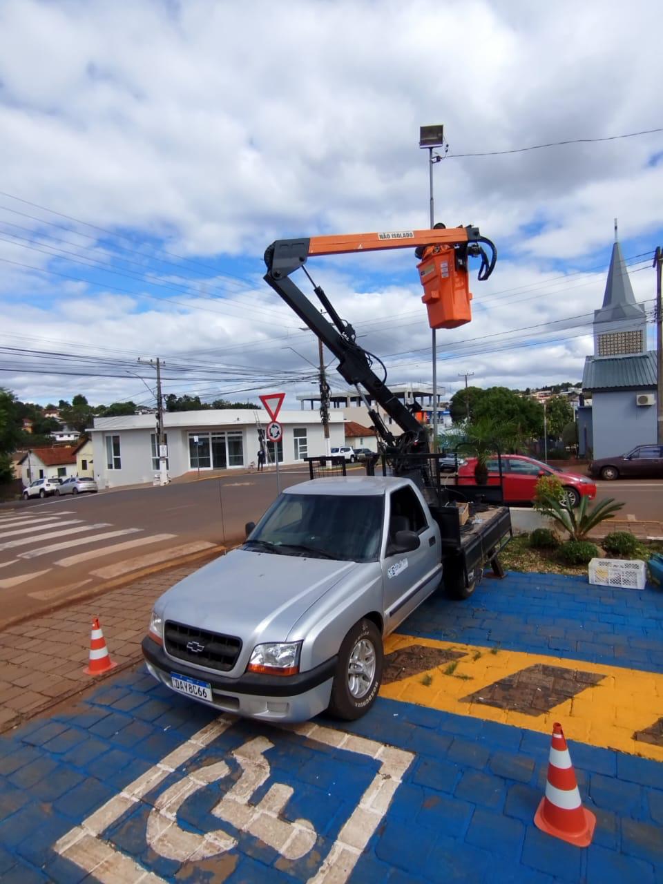 Caminhonete prata com cesto aéreo laranja estacionada em praça com cones de trânsito
